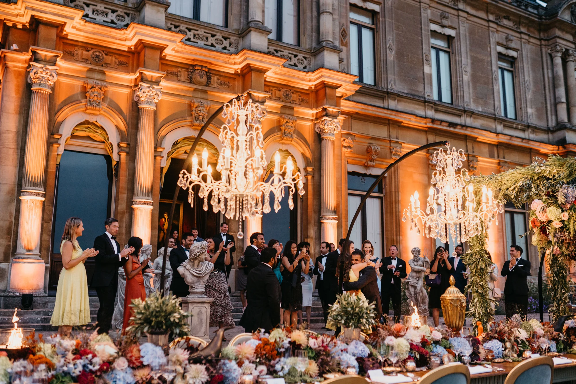 Guests standing in front of the uplit facade before taking their seats, flowers massed along the table edge.