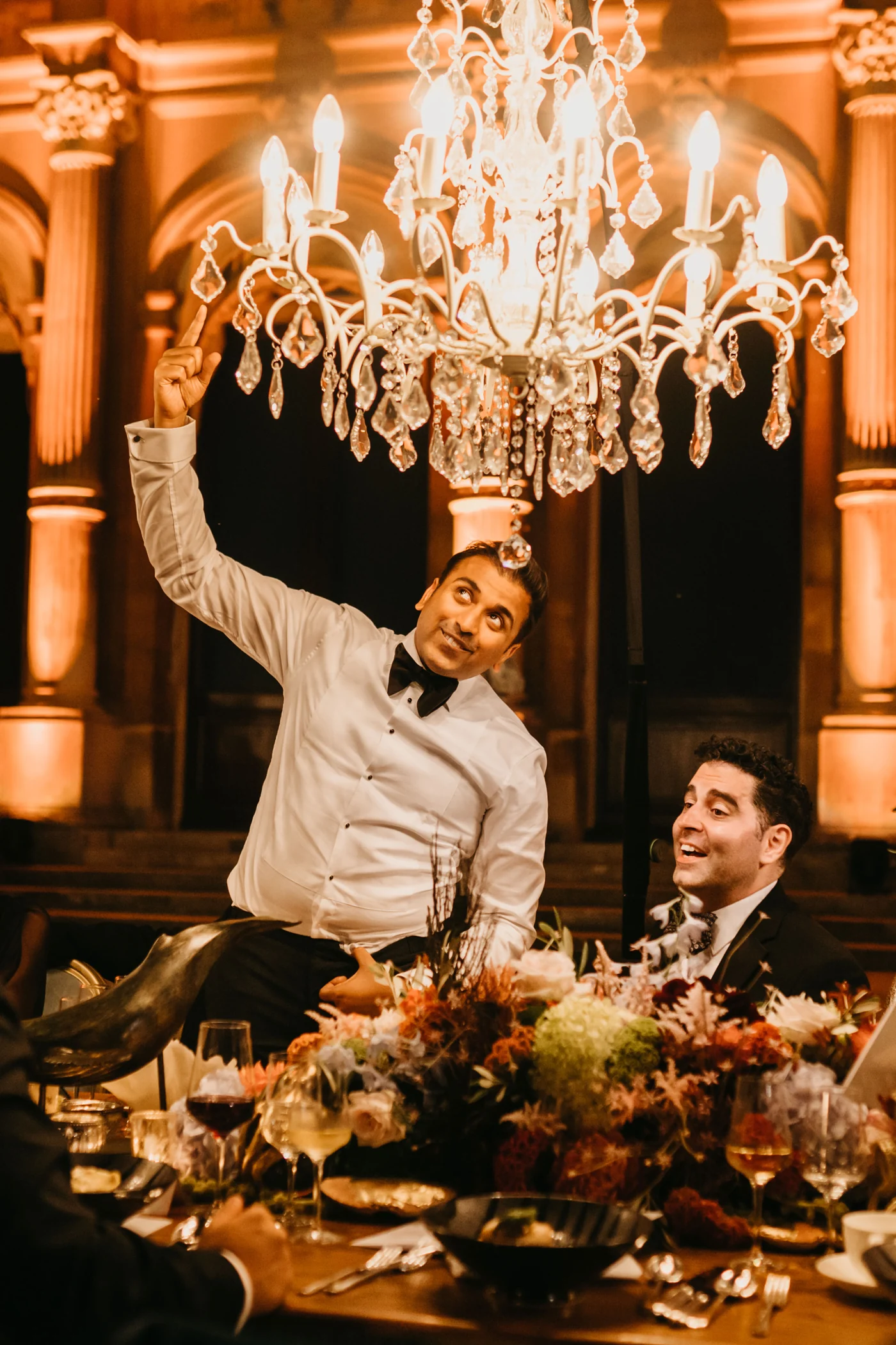 A guest in white throwing his arms up at the table, a crystal chandelier lit above him.