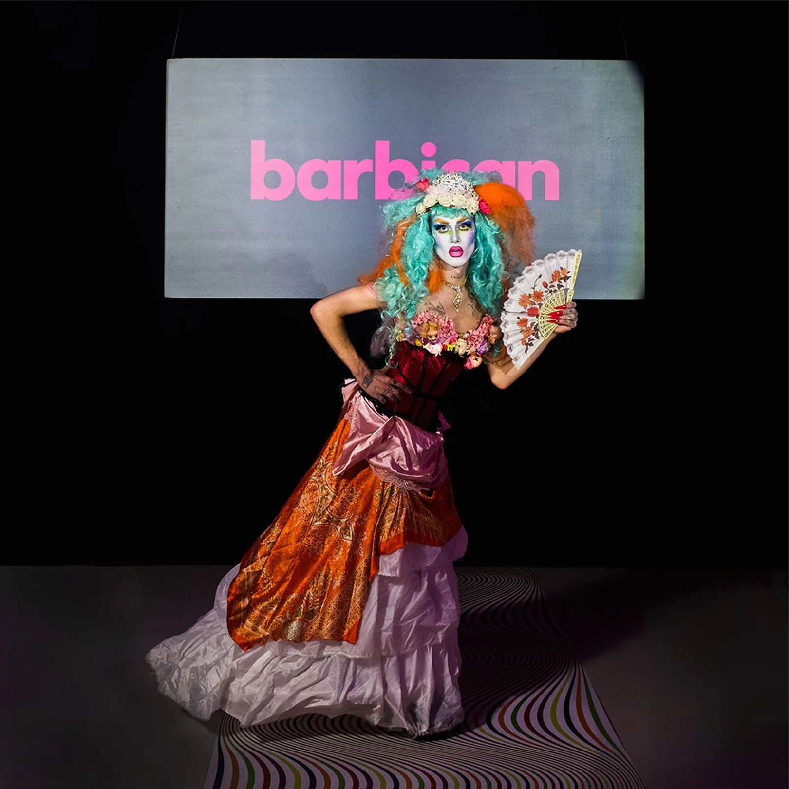 A performer in period costume and blue-green wig posing in front of a lit Barbican screen.