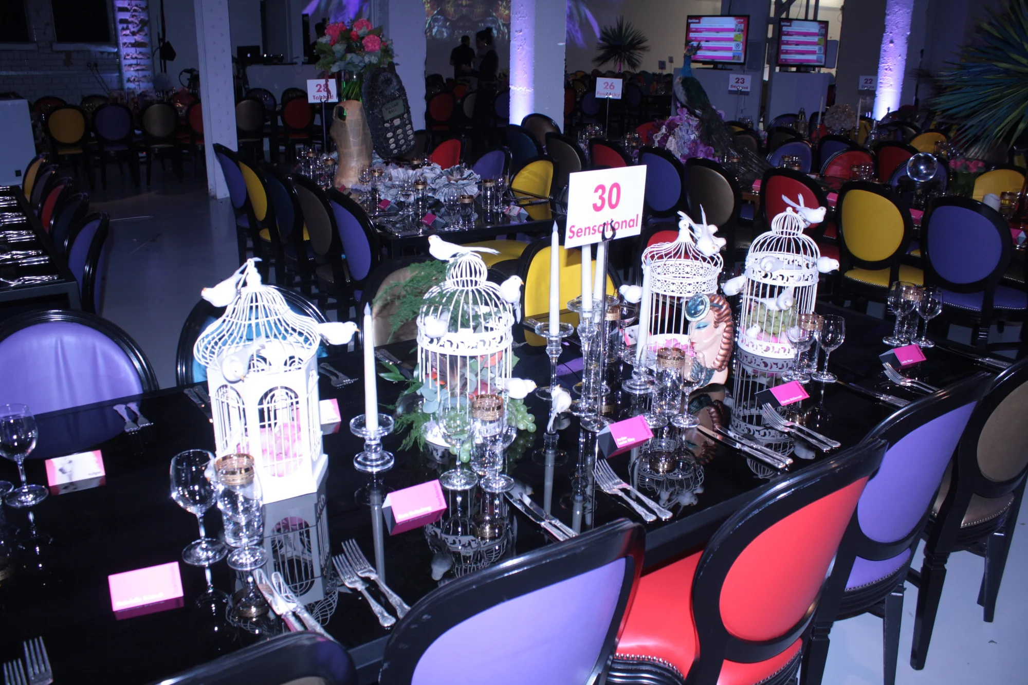 A dinner table dressed with white birdcages and mismatched coloured chairs down both sides.