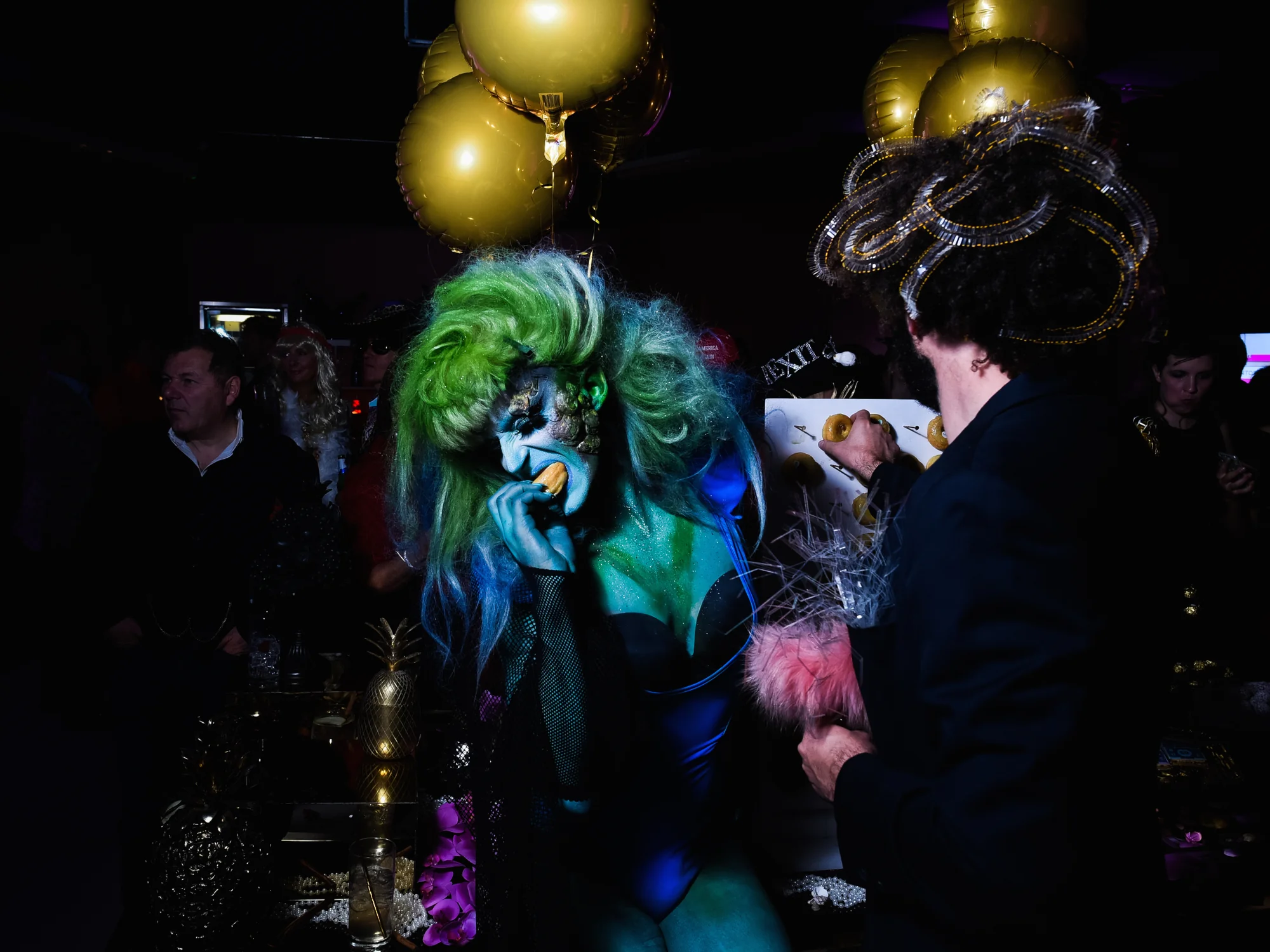 Gold balloons massed above the guests, a headdress of green hair below them.