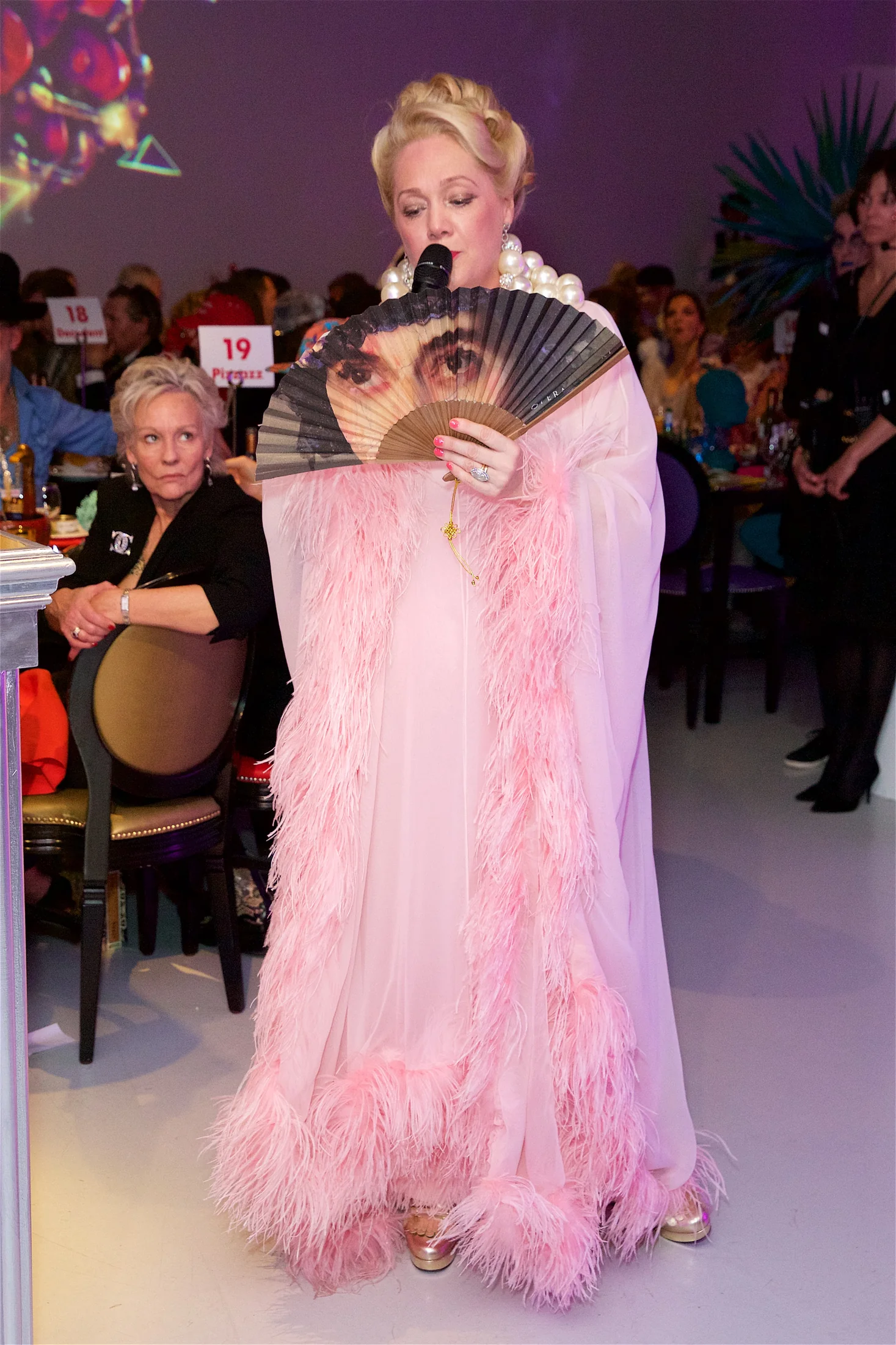 A guest in a pink feathered gown holding a printed face fan in front of her.