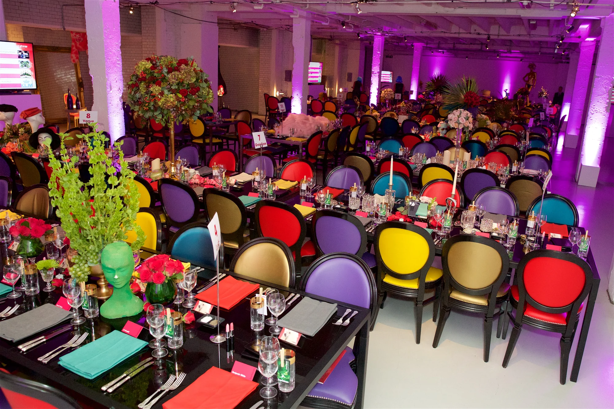The full spread of dressed tables under pink light, every chair a different colour and every table differently themed.