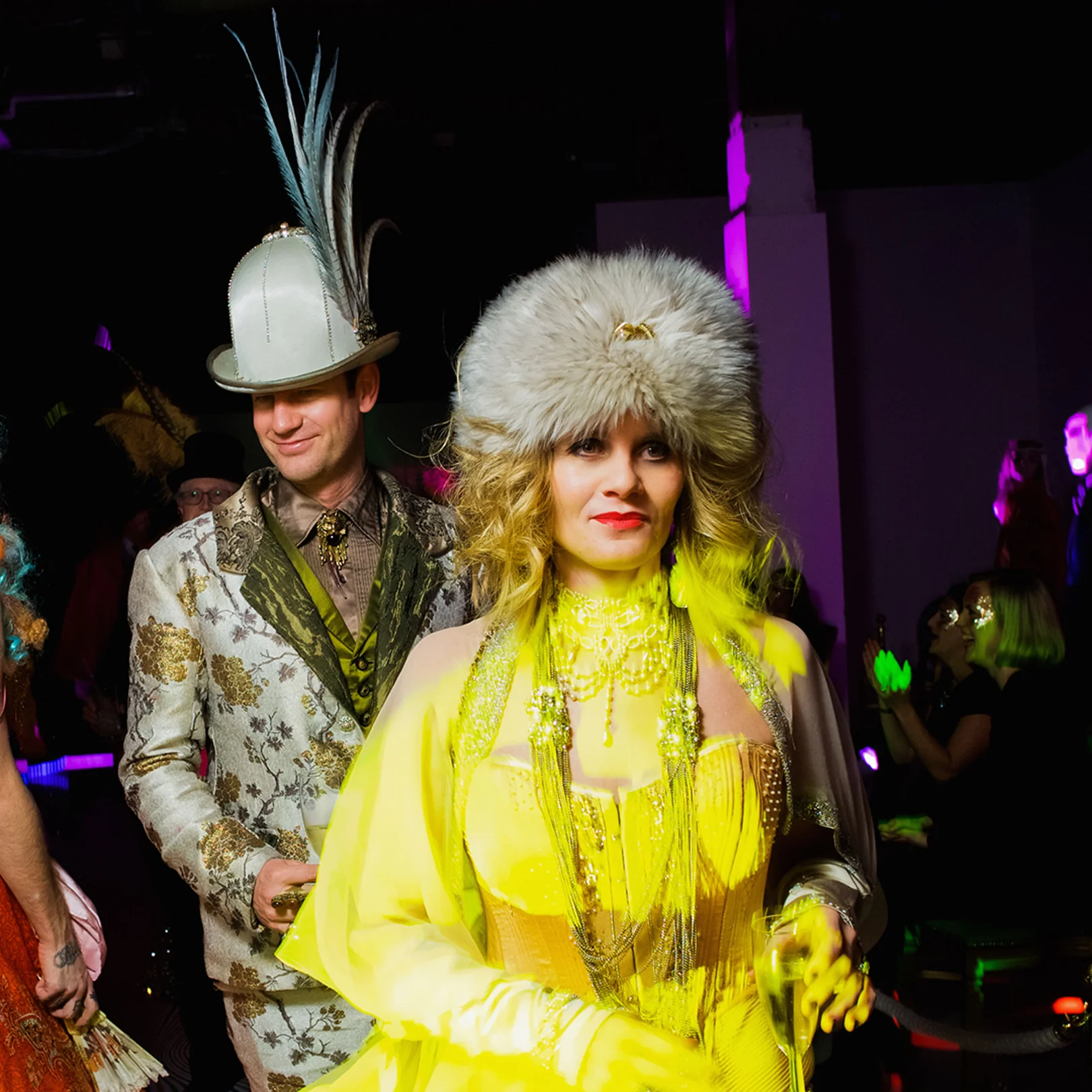A guest in a yellow fringed gown and fur hat walking past the tables with another in brocade.