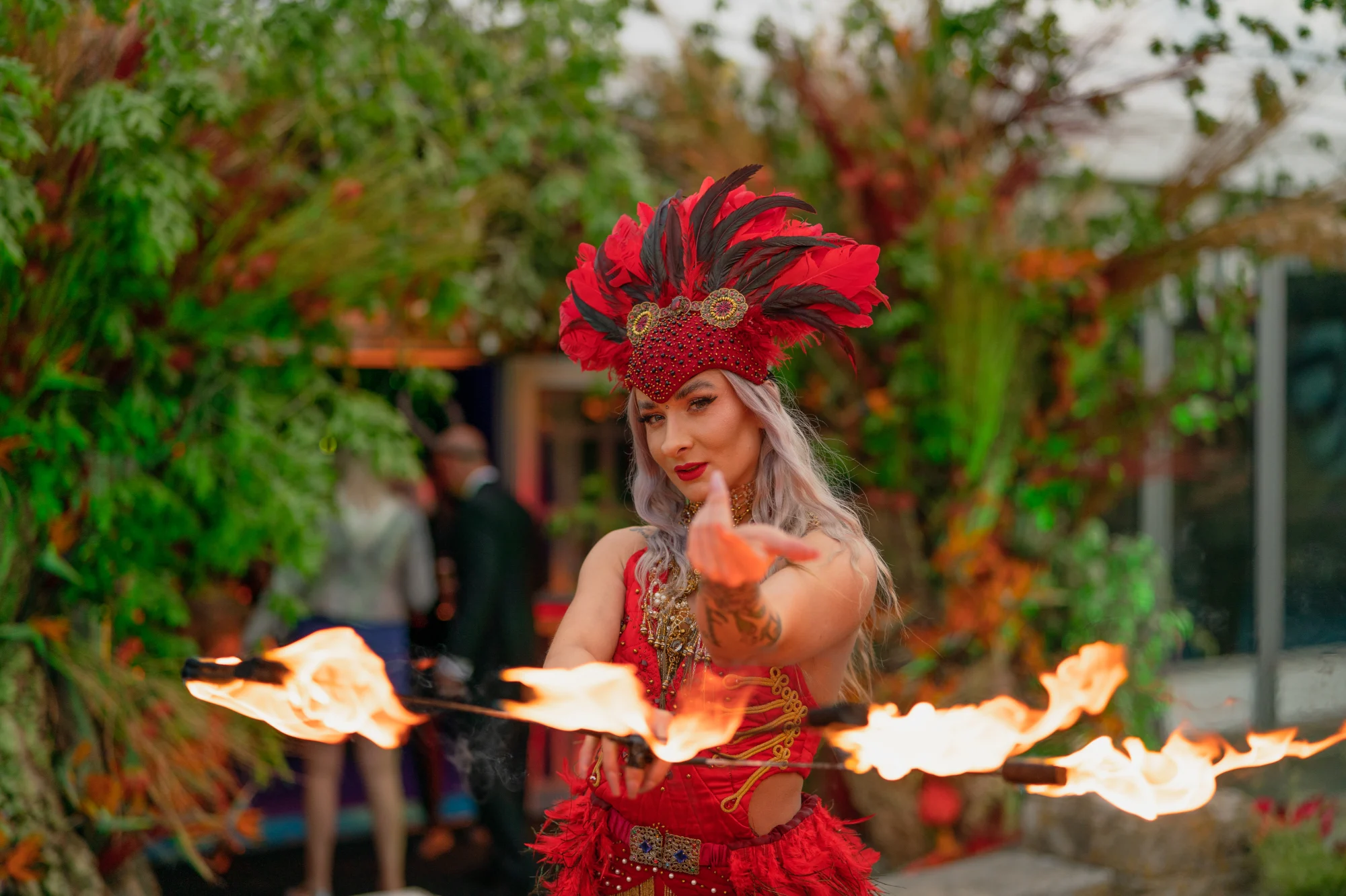 A performer in a red and black feathered headdress and beaded costume spinning two lit fire staves, greenery and guests behind her.