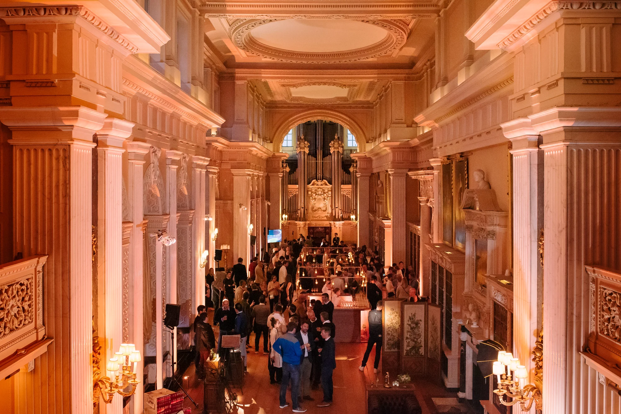 Looking down the length of a palace library from a balcony, fluted columns and a carved ceiling above, an organ at the far end and guests gathered around a lit bar below.