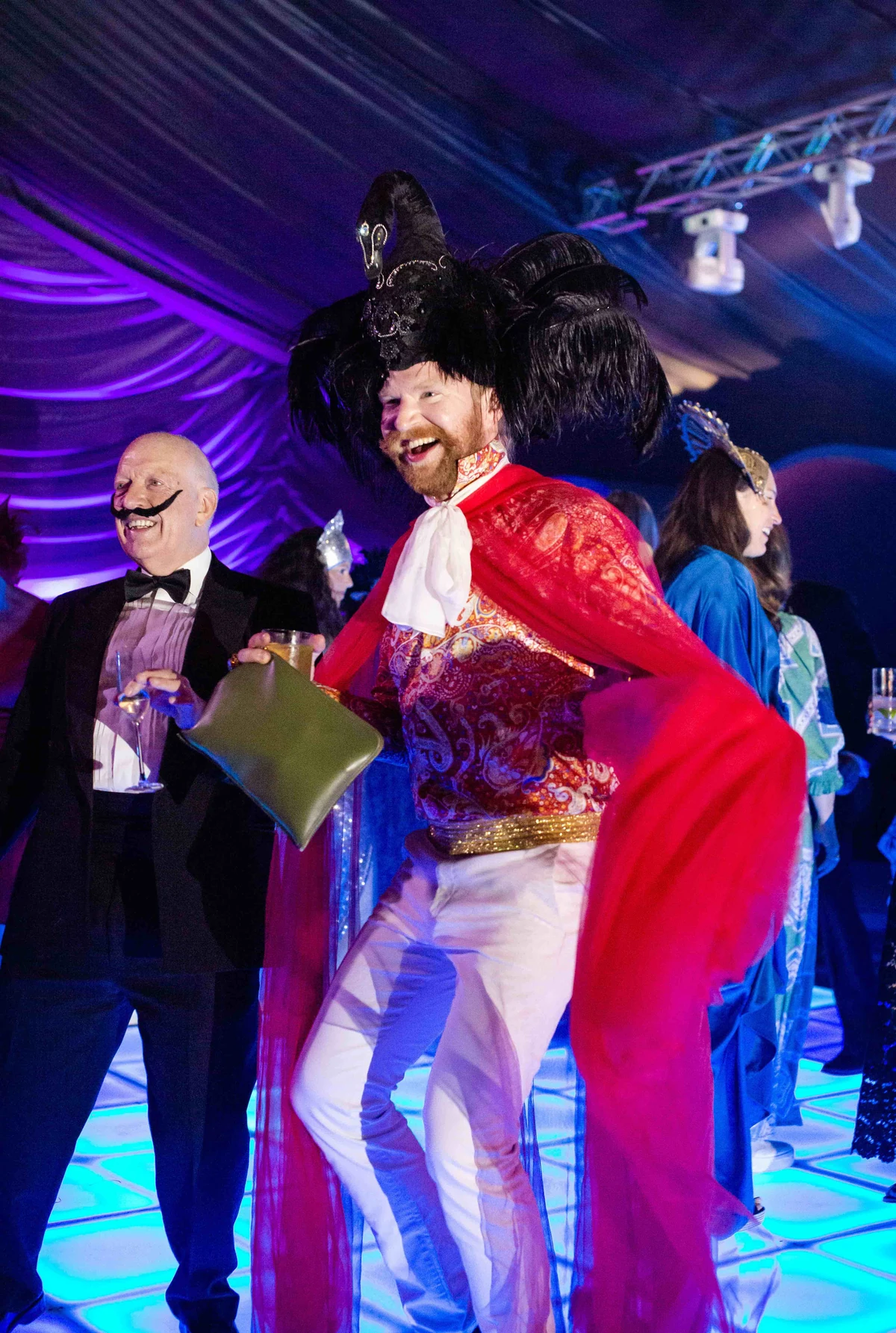 A performer in a red cape, embroidered corset and towering black plume, laughing beside a man in a monocle, under purple and blue light.