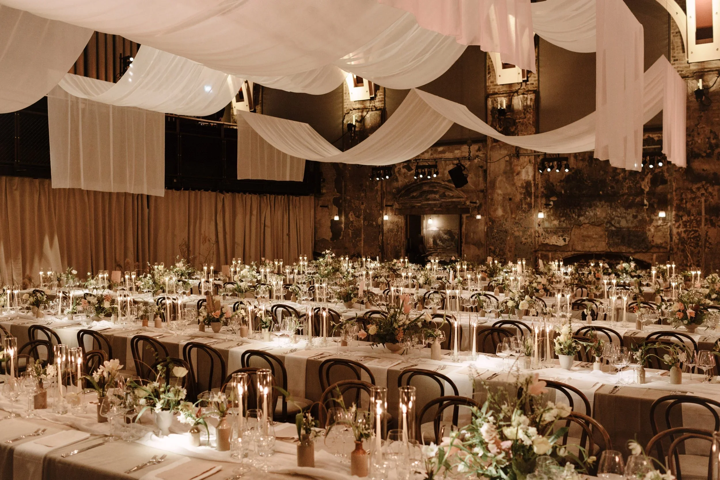 A hall at Battersea Arts Centre dressed for a gala, pale drapes swagged beneath the ceiling and long tables set with candlelight and flowers.