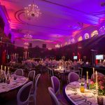 A Winter Wassail for War Child: round tables laid with candles and flowers under a ceiling washed purple, the stage lit at the far end.