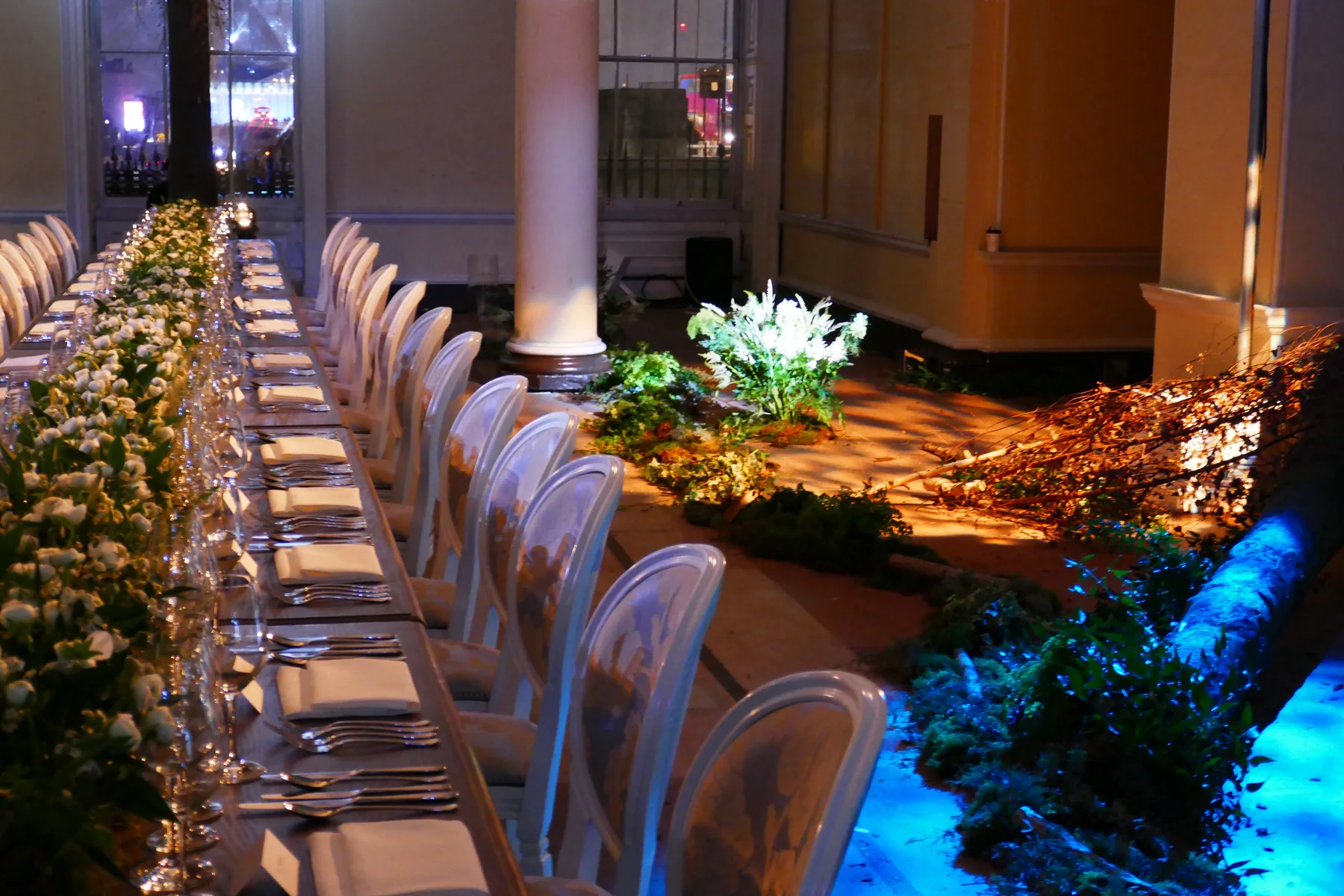 A long banqueting table laid with white florals and glassware running its length, beneath a colonnade with lit planting alongside.