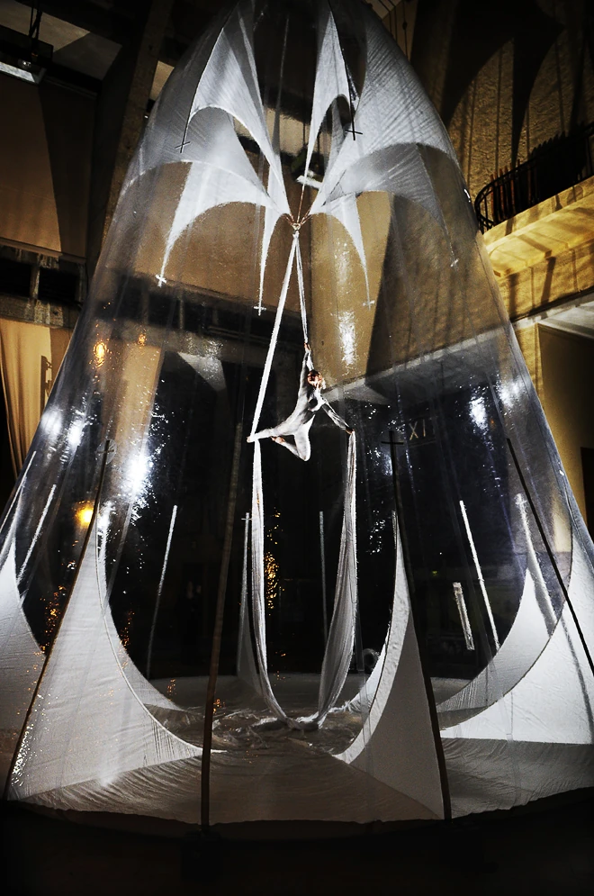 An aerialist inside a tall cone of white fabric suspended from the truss in Lindley Hall, lit from within.