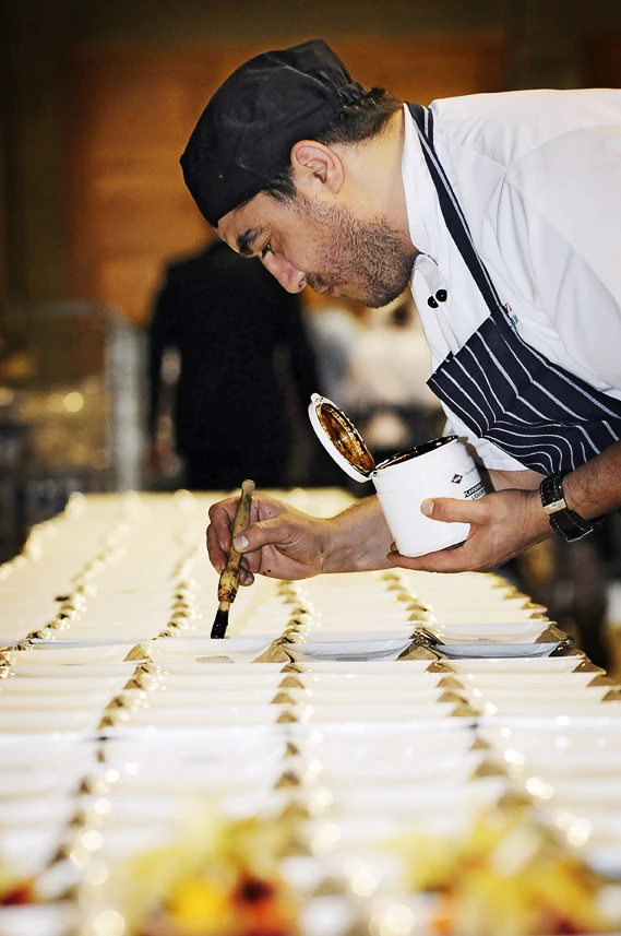 A chef in whites and a striped apron bent over a long table of plated starters, finishing each one with a sauce from a small pot.