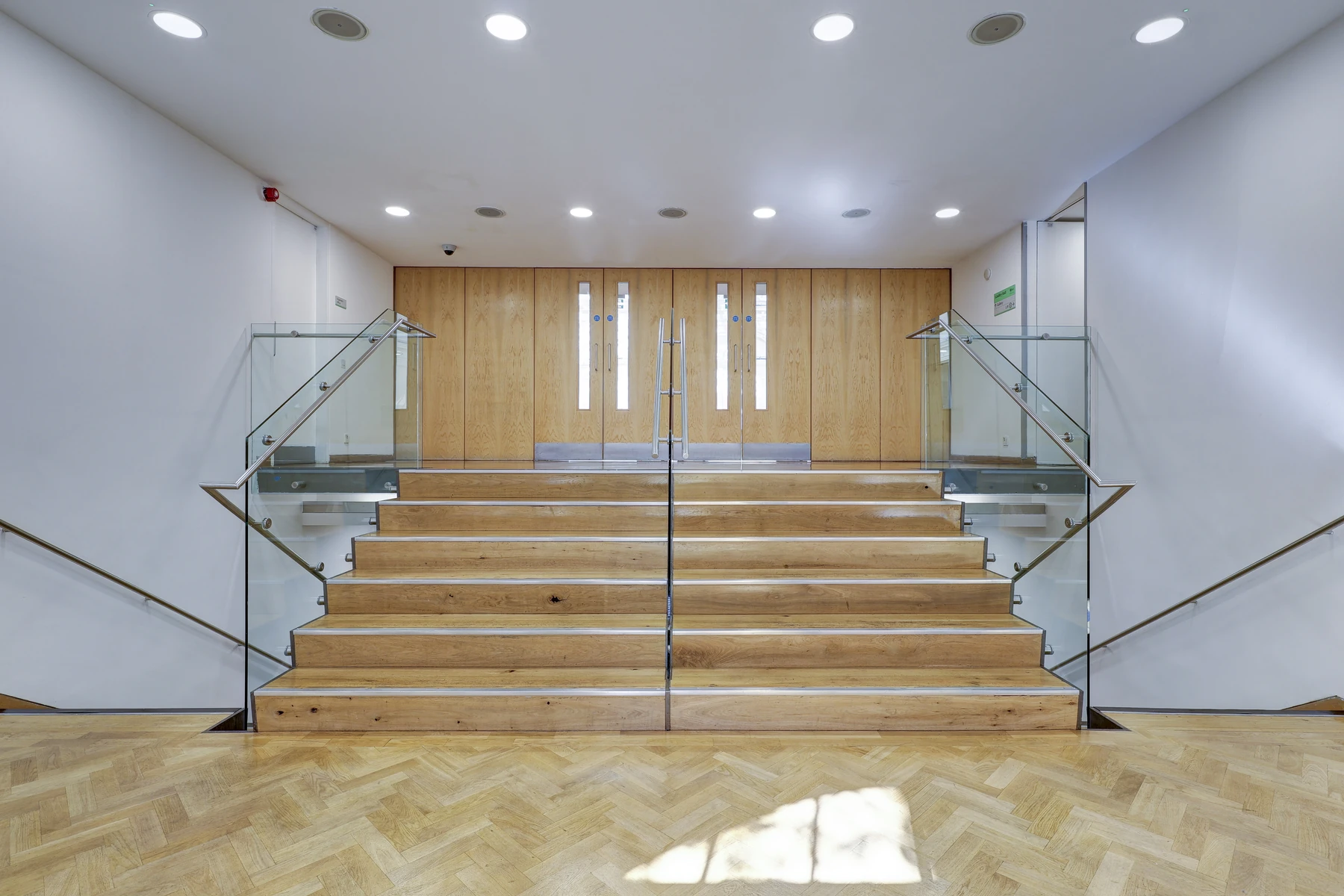 The foyer at Lindley Hall: a wide flight of oak steps with glass balustrades rising to a pair of oak doors, parquet floor in the foreground.