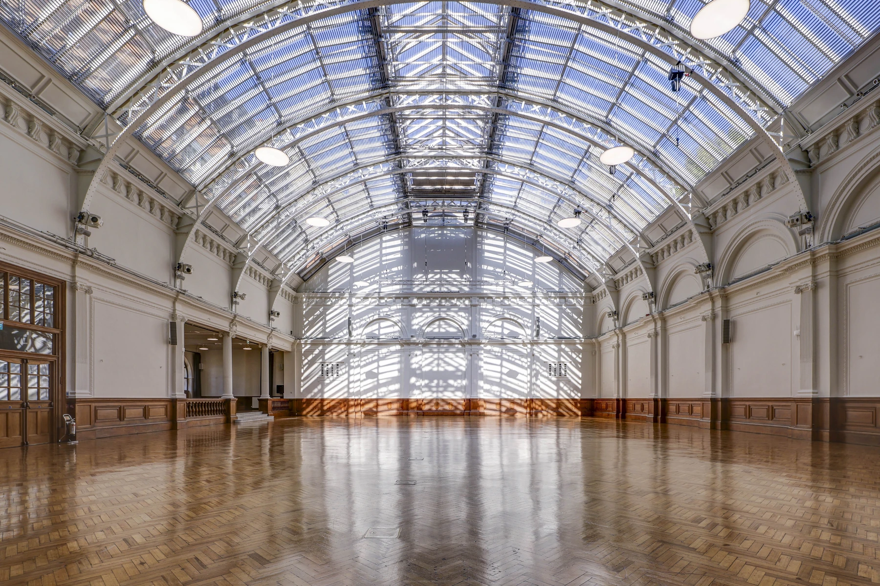 The Main Hall of Lindley Hall empty and in daylight, seen from the entrance: the glass and iron barrel vault overhead, white walls and arcades either side and the parquet floor running to the glazed far end.