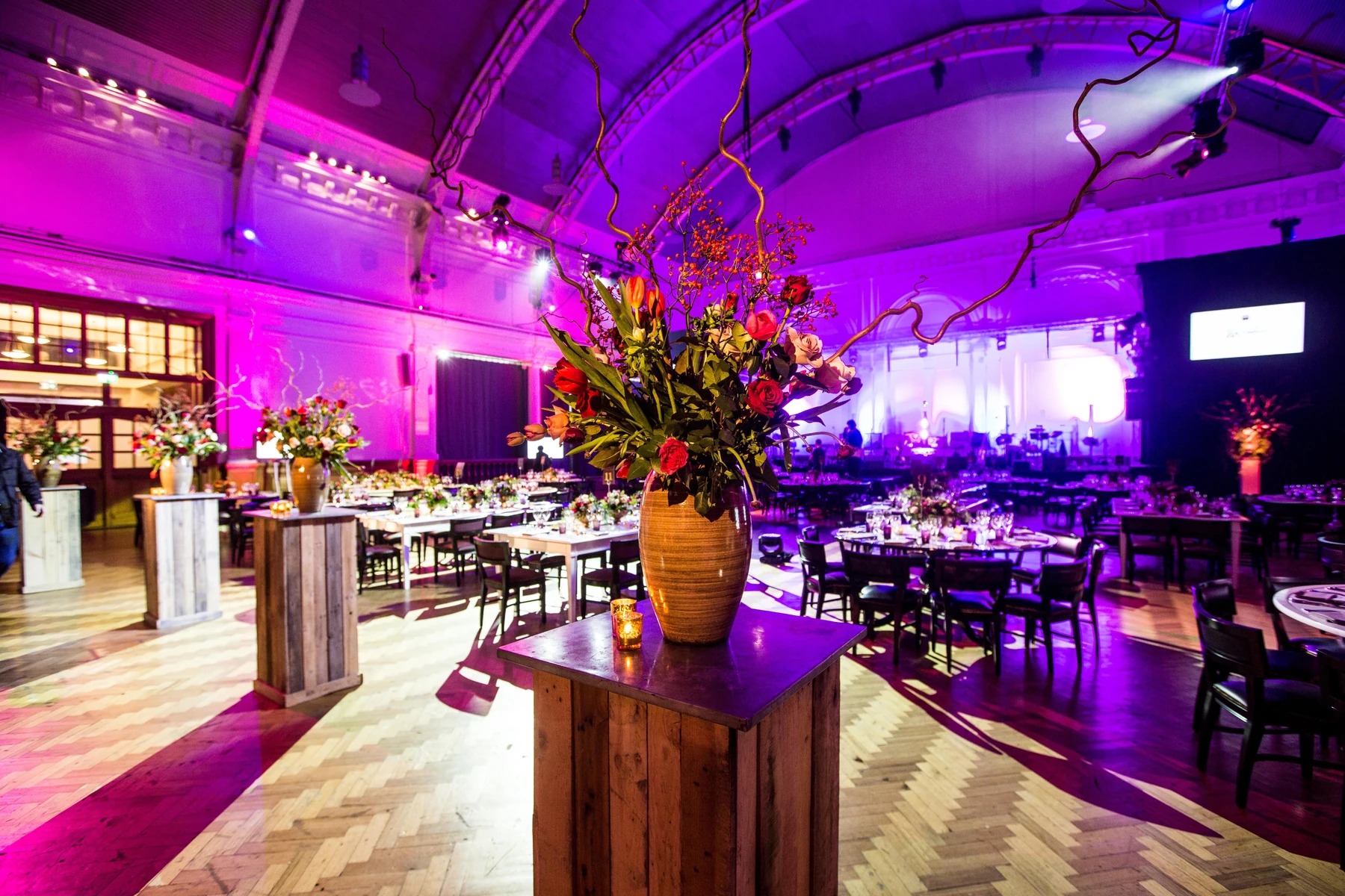 Lindley Hall laid for a gala, seen from beside the bar: the glass vault lit purple, tall flowers on wooden plinths, round tables with place settings, the stage and screens at the far end.
