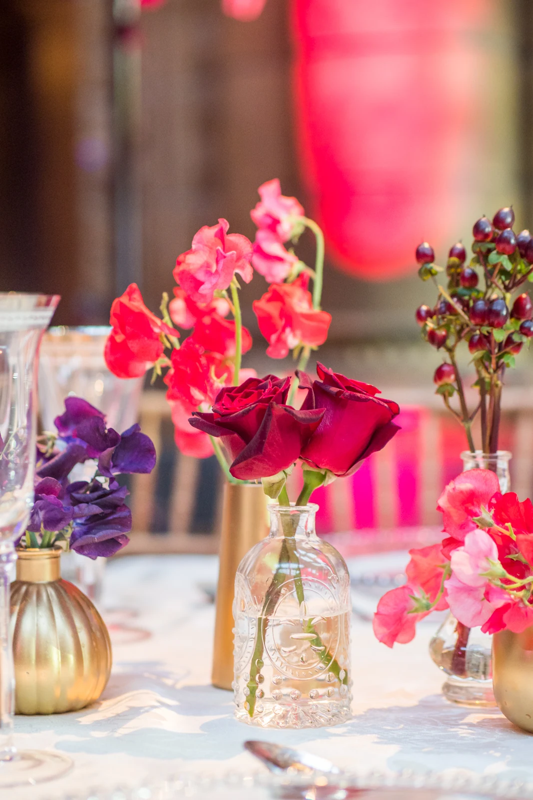 Gold and clear glass bud vases of red roses, pink sweet peas and purple flowers on a cream tablecloth, lit pink.