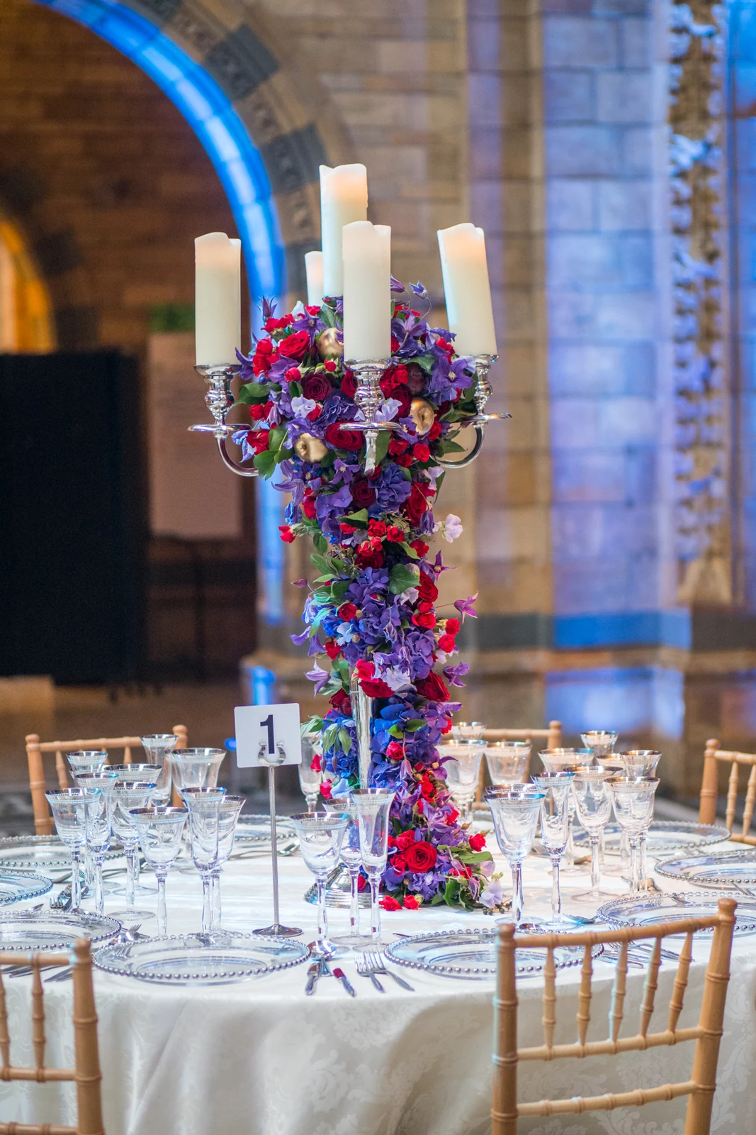 A tall candelabra wrapped in red roses and purple flowers on a round table, five white candles at the top, an arch of the hall lit blue behind.