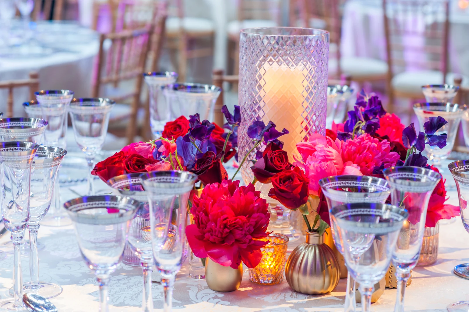 Red roses and peonies in gold vases beside a cut-glass hurricane lamp and gold-rimmed glassware on a cream cloth.