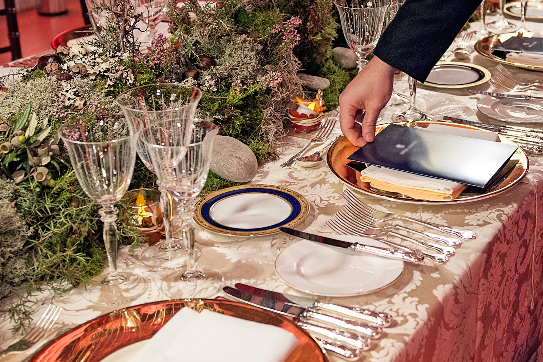 A place setting close, a hand placing the menu card on a copper charger.