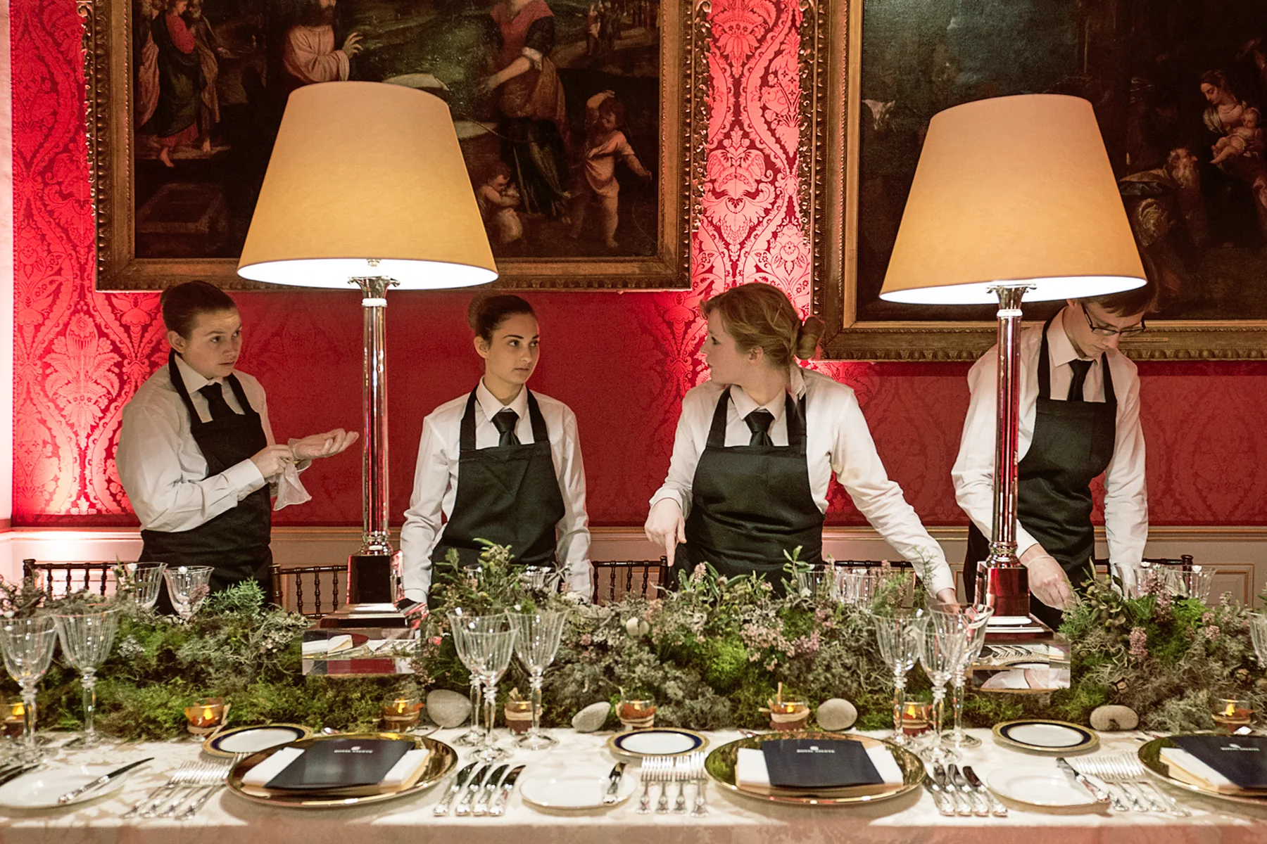 Four staff standing to the laid table, lamps lit above the foraged centre.