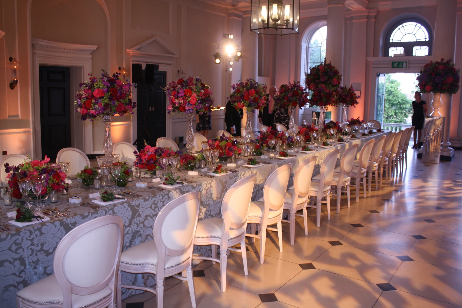 Seamen’s Hall laid for one long table, white chairs down both sides, tall arrangements of roses on glass stands, the columns lit pink and the chequered marble floor.
