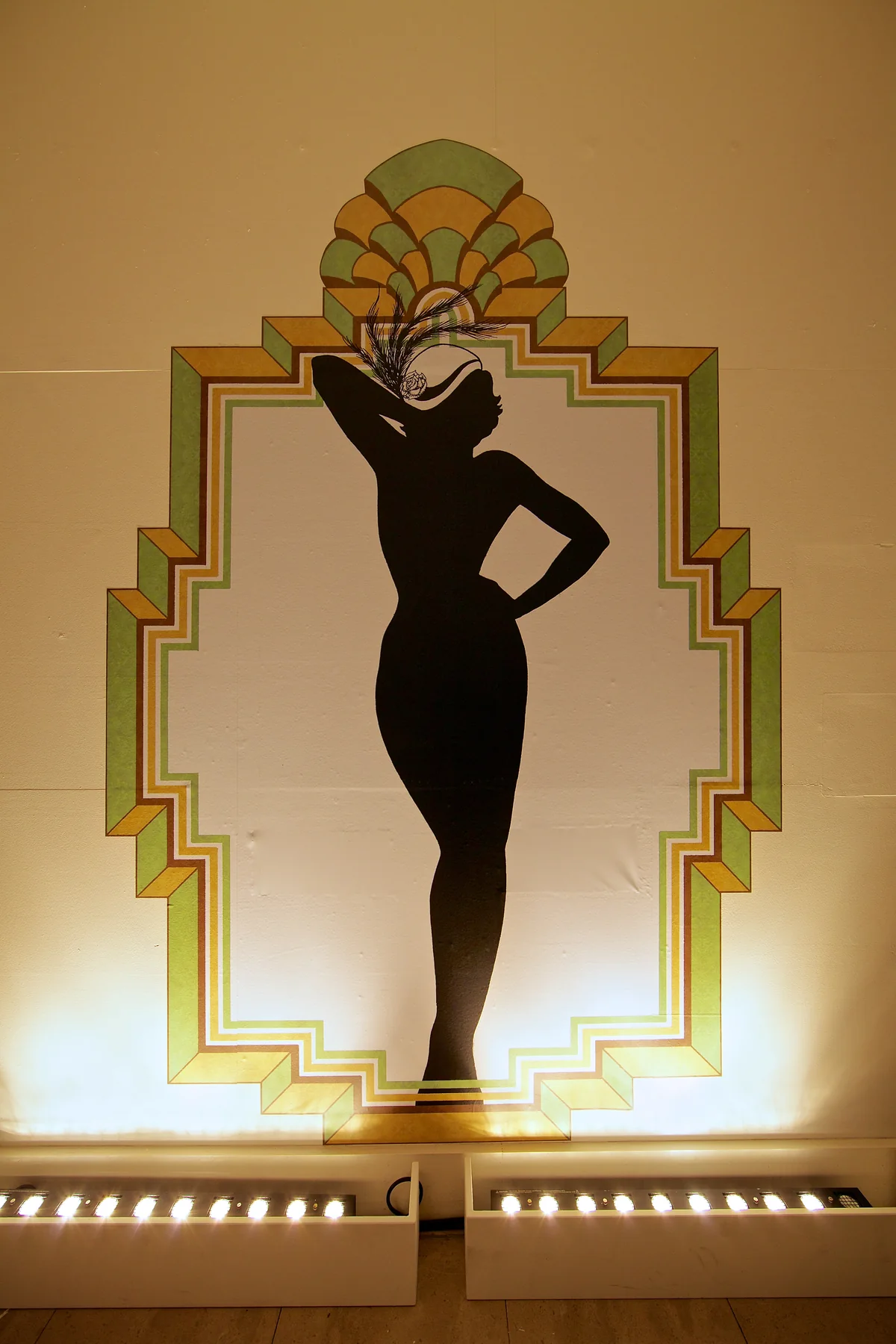A cabaret dancer in silhouette, hand on hip beneath a feathered fan headdress, inside a stepped Art Deco frame.