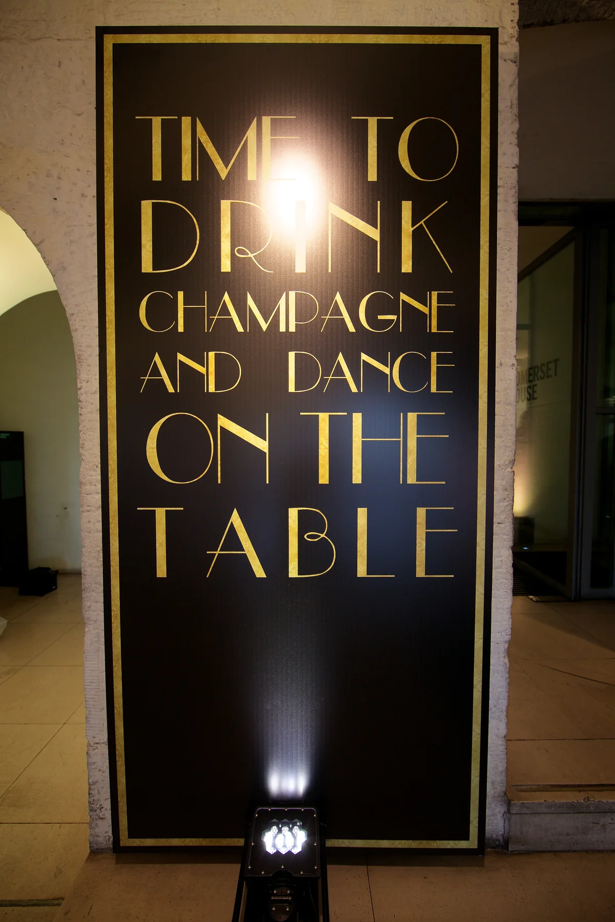 A gold on black sign reading TIME TO DRINK CHAMPAGNE AND DANCE ON THE TABLE.