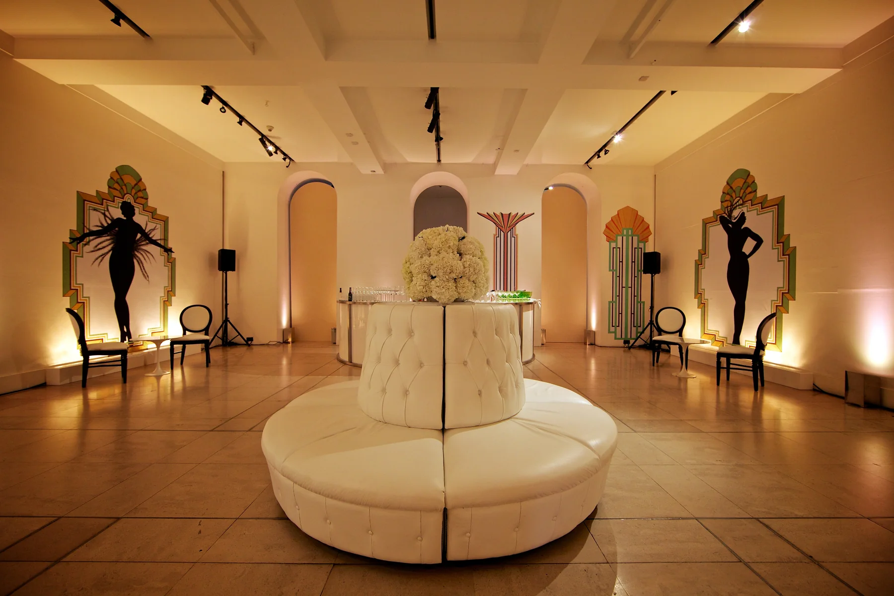 The finished space at Somerset House, a circular cream banquette beneath a white hydrangea ball, cabaret dancer panels lit along the walls.